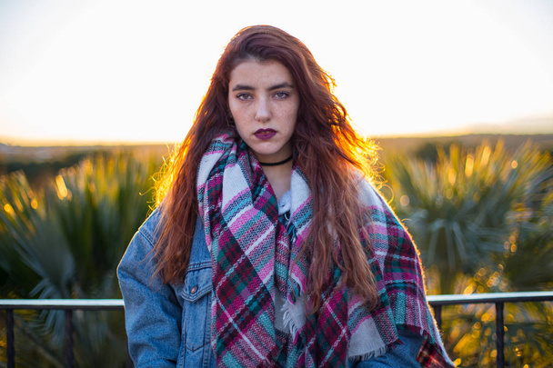 Young brunette in trendy denim jacket and scarf looking alluringly at camera in back lit on nature.  - Photo, Image
