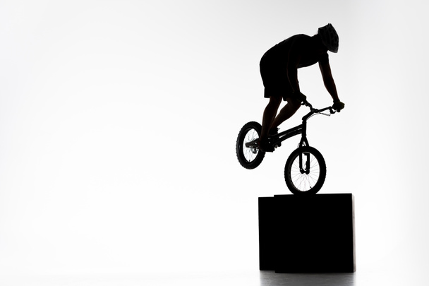 silhouette of trial cyclist performing stunt while balancing on cube on white - Photo, Image