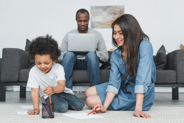 Afro-Amerikaanse moeder en zoon puttend uit vloer thuis, vader werken met laptop  - Foto, afbeelding