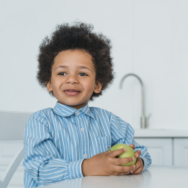 adorable african american boy holding apple at table in kitchen - Φωτογραφία, εικόνα