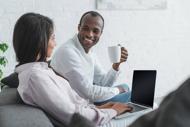 african american girlfriend using laptop and boyfriend drinking coffee at home - Photo, Image