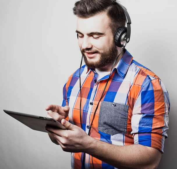Studio πορτρέτο ενός όμορφου γενειοφόρου άνδρα χρησιμοποιώντας ένα tablet με ακουστικά πάνω από ένα ανοιχτό γκρι φόντο. - Φωτογραφία, εικόνα