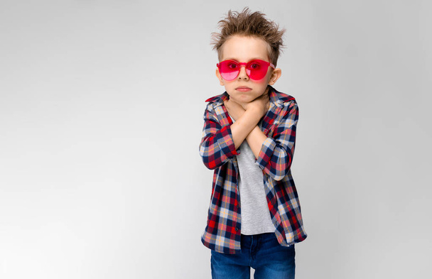 Nice caucasian preschooler boy in casual outfit and red sunglasses showing different expressions on white wall in studio. Holding neck. - Photo, Image
