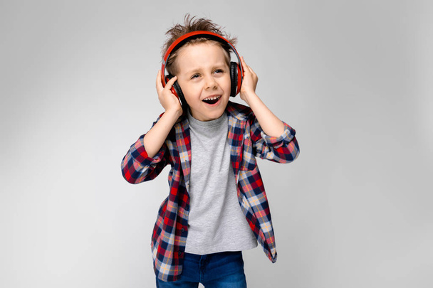 Niño preescolar caucásico agradable en traje casual posando con auriculares rojos y mostrando diferentes expresiones en la pared blanca en el estudio
. - Foto, imagen