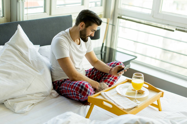 Junger Mann frühstückt im Bett, während er sein Handy benutzt - Foto, Bild