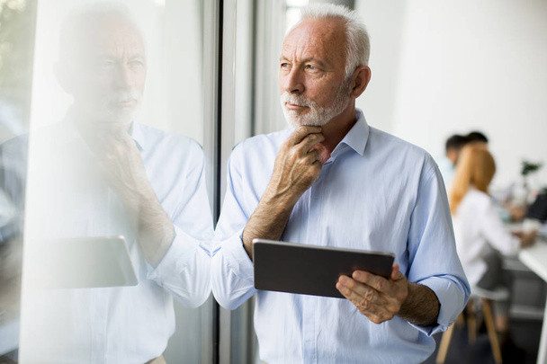 Portret van senior zakenman permanent door raam met digitale tablet in zijn hand - Foto, afbeelding