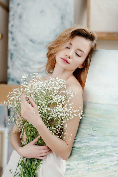 sinnliche nackte Mädchen mit geschlossenen Augen mit weißen Blumen und lehnt an Staffelei im Kunststudio - Foto, Bild