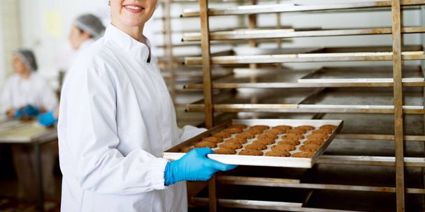jovem trabalhador feminino feliz segurando biscoitos recém-assados em folha de flandres na fábrica de produção de alimentos
 - Foto, Imagem