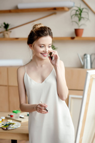 schöne lächelnde junge Malerin mit Pinsel, während sie mit dem Smartphone spricht und im Kunstatelier auf die Staffelei blickt - Foto, Bild