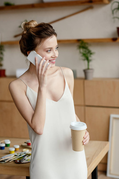 beautiful smiling young woman holding coffee to go and talking by smartphone in art studio - Photo, Image