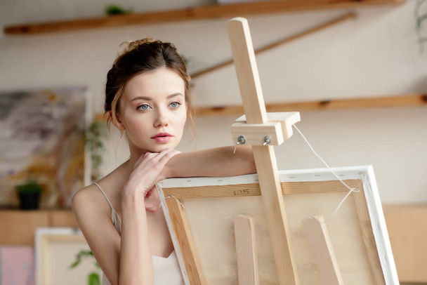 schöne nachdenkliche junge Künstlerin lehnt an Staffelei und schaut weg - Foto, Bild