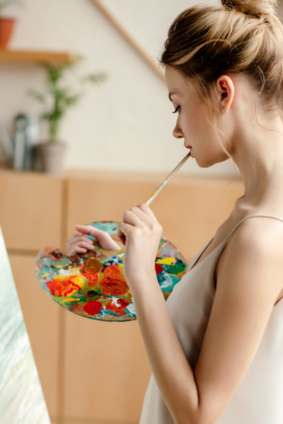 side view of attractive young artist with palette and brush painting on easel in studio - Photo, Image