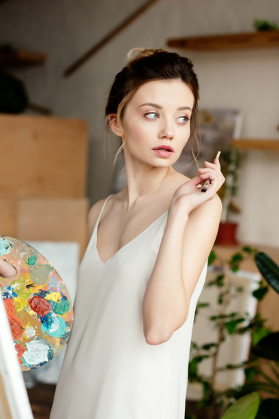 beautiful young artist holding palette and looking away in studio - Foto, Imagem