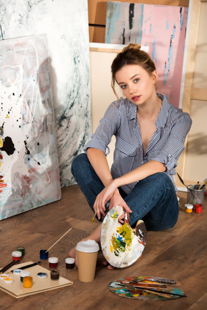 piękna Młoda Artystka siedzi na podłodze i patrząc na kamery w studio sztuki - Zdjęcie, obraz