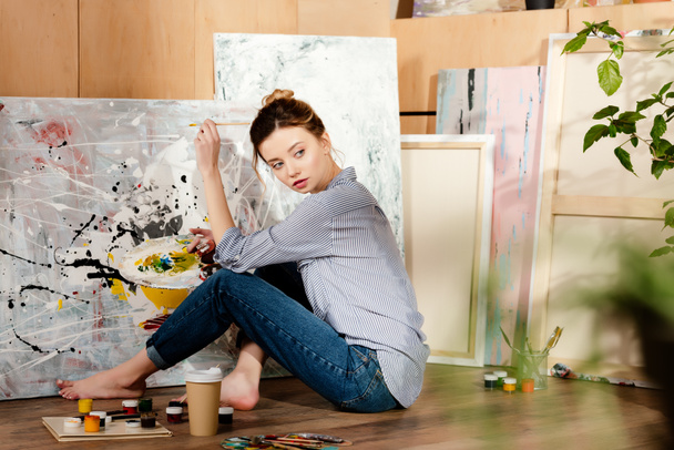 beautiful young barefoot artist sitting on floor and painting picture in studio - Photo, Image