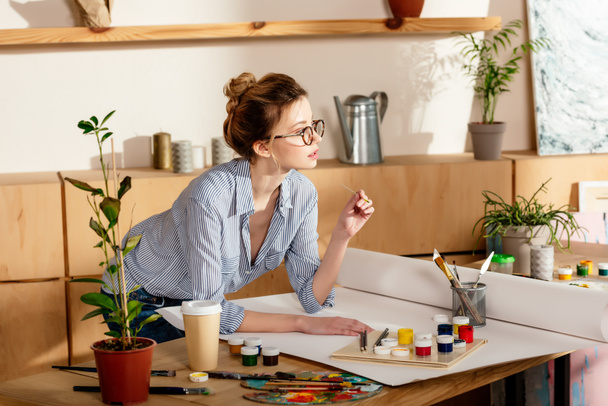 joven artista femenina en gafas de pie en la mesa con lienzo vacío y materiales de pintura
  - Foto, imagen