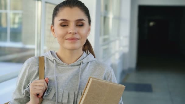 Portrait of young beautiful caucasian confident female student standing in white glassy corridor smiling positively holding textbook looking at camera - Video