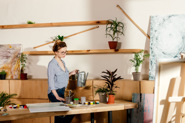 junge stilvolle Künstlerin in Brille gießt Topfplan im Atelier - Foto, Bild