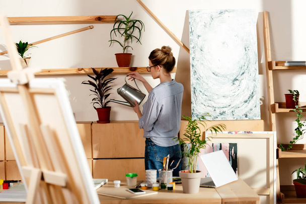 visão traseira do artista feminino regando plano em vaso no estúdio
  - Foto, Imagem
