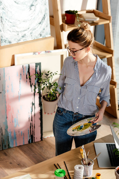 stylish female artist in eyeglasses holding plant and palette in studio  - Foto, afbeelding