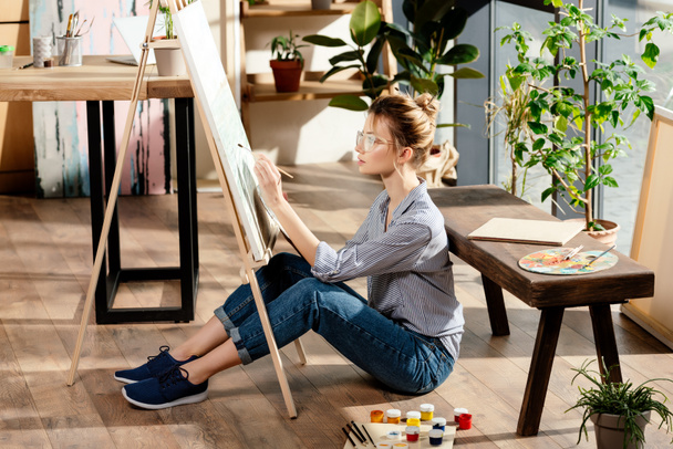 young stylish female artist in eyeglasses painting on easel  - 写真・画像