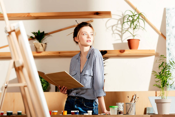joven atractiva artista femenina sosteniendo libro y mirando hacia otro lado
  - Foto, Imagen