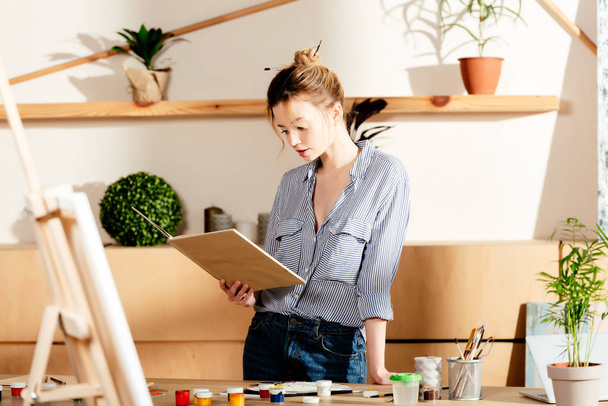 nuori naistaiteilija lukee kirjaa ja seisoo pöydässä maalaustarvikkeiden kanssa
  - Valokuva, kuva