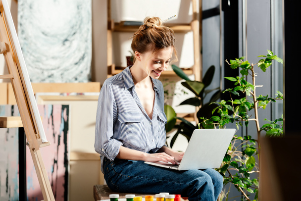sorrindo jovem mulher digitando no laptop e sentado no banco com tintas
 - Foto, Imagem
