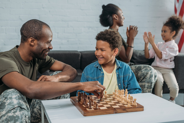 Χαμογελαστός στρατιώτης παίζει σκάκι με το γιο του ενώ η μητέρα και κόρη, παίζουν μαζί - Φωτογραφία, εικόνα