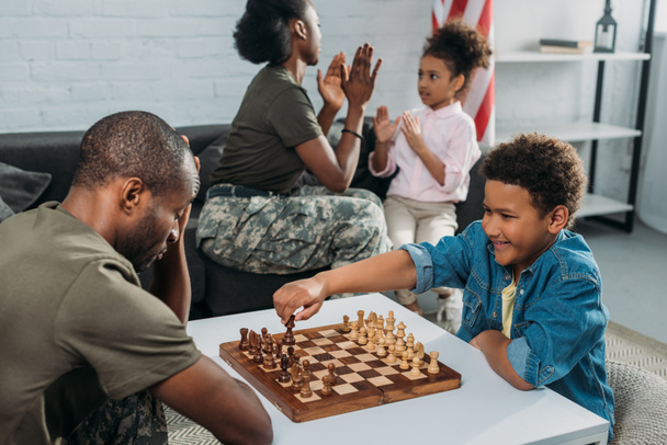 Padre en uniforme del ejército jugando ajedrez con su hijo mientras madre e hija juegan juntas
 - Foto, Imagen