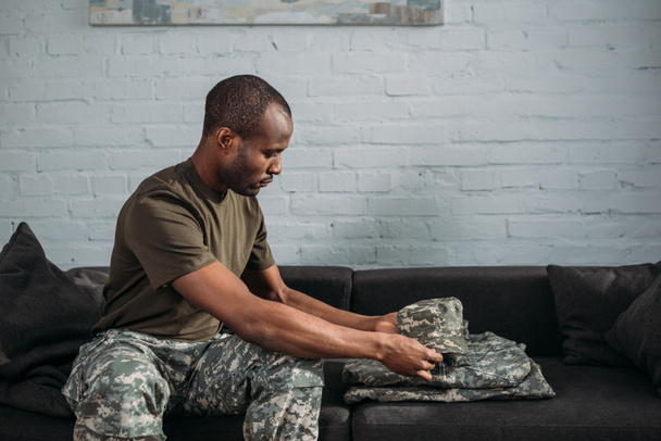 African american male soldier folding camouflage clothes on sofa - Photo, Image