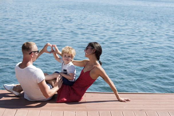 Mother and father making heart or love gesture with hands near their child. Happy family spend time together, sea background. Parents with son sit on seafront, rear view. Family vacation concept. - Photo, image