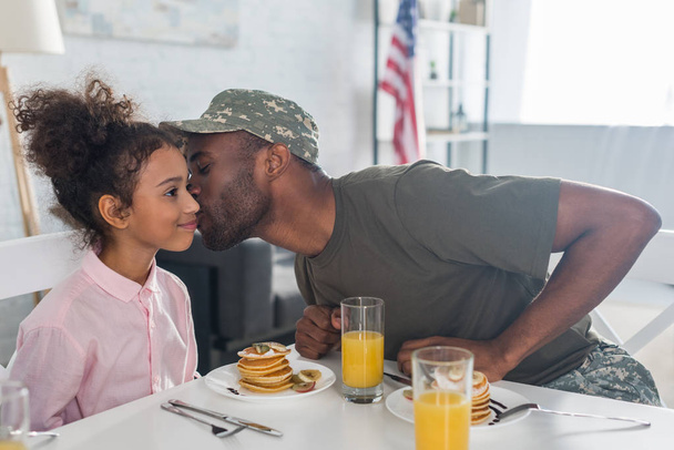 Πατέρα του στρατού ομοιόμορφο φιλιά αφρικανική αμερικανική κόρη από το τραπέζι της κουζίνας - Φωτογραφία, εικόνα