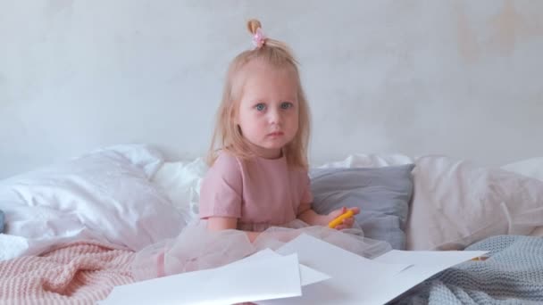 Little charming blond girl in pink dress holding a felt-pens and sitting on bed among paper. - Video