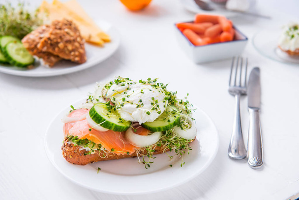 Cereal bun with egg benedict, smoked salmon, sprout micro greens, onion and cucumber slices, cream cheese on the served white wooden table. Healthy breakfast concept. Selective focus. - 写真・画像