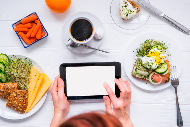 Close up female hands holding tablet with blank copy space screen and having healthy breakfast and coffee. Work, planning, edication during eating. Selective focus, top view. - Photo, image