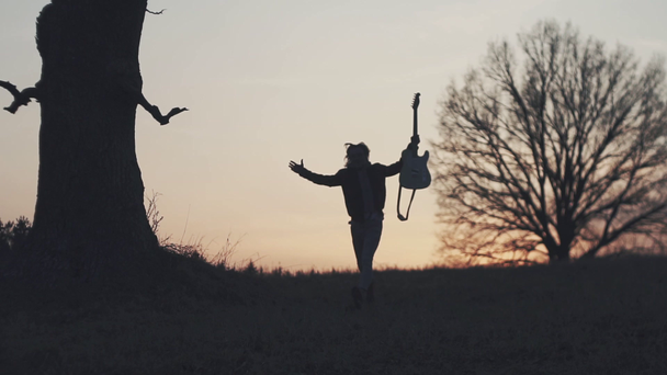 Σιλουέτα του κιθαρίστα στο ηλιοβασίλεμα αργή κίνηση. τρέχει με μια κιθάρα στα χέρια του - Πλάνα, βίντεο