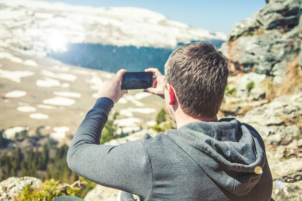 travel guy sits on top of the mountain and takes pictures of the smartphone View from back of the tourist traveler on background mountain - Foto, immagini