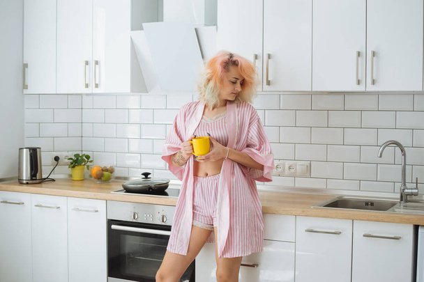 Menina bonita nova com cabelo rosa em pijama rosa bebendo chá enquanto está de pé na cozinha loft branco
 - Foto, Imagem