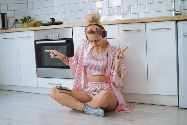 Happy young woman with pink hair dancing in kitchen wearing pink pajamas and listening to music with headphones - Foto, imagen