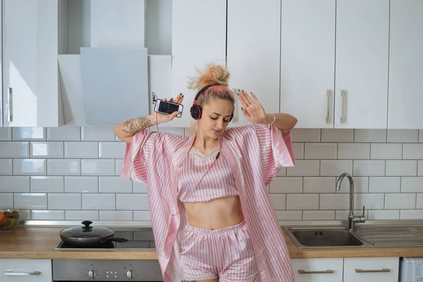 Mujer joven y feliz con el pelo rosa bailando en la cocina con pijama rosa y escuchando música con auriculares
 - Foto, imagen