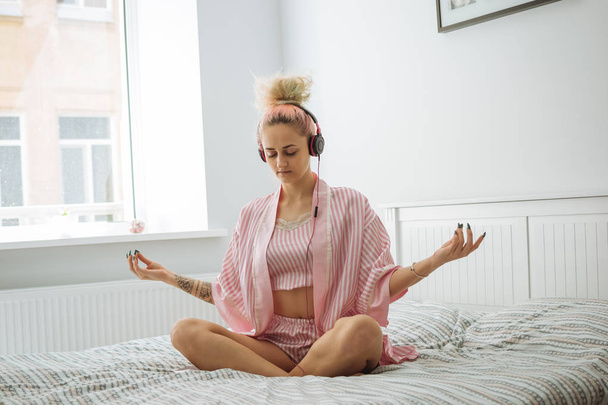 Young woman with pink hair in pink pajamas meditating with headphones - Photo, Image