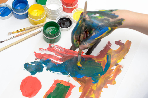 Το χέρι ενός παιδιού στη ζωγραφική χρώματα γκουάς σε χαρτί λευκό. - Φωτογραφία, εικόνα
