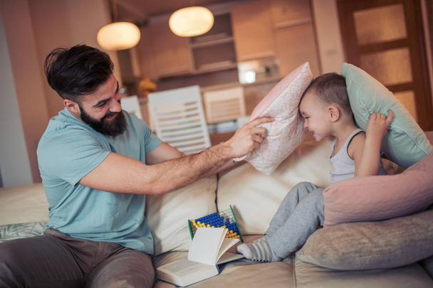 Kleine jongen en vader vechten met kussens en glimlachen terwijl zittend op bed thuis. - Foto, afbeelding