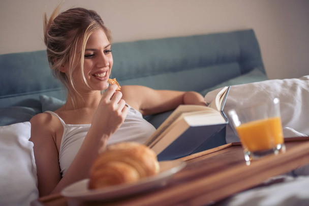 Onnellinen nuori nainen lukee kirjaa ja syö sängyssä aamulla
. - Valokuva, kuva