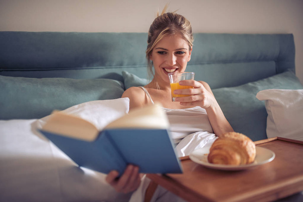 Happy young woman reading book and drinking juice in bed in morning. - Foto, Imagen