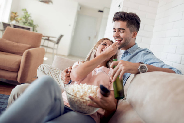 若いカップルがソファの上に横たわって、映画を見て、ポップコーンを食べて. - 写真・画像