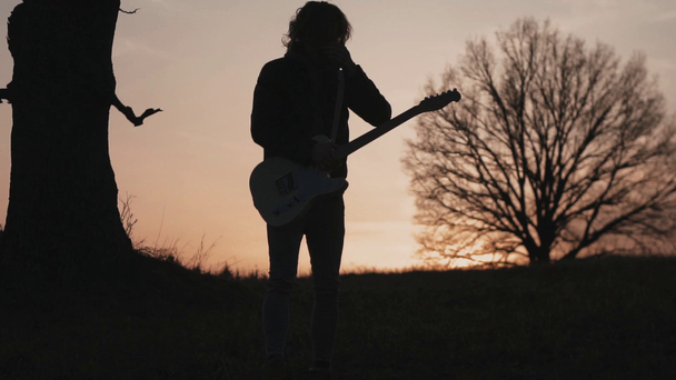 man plays electric guitar and sings a lyric song in a field near the tree at sunset. silhouette - Footage, Video