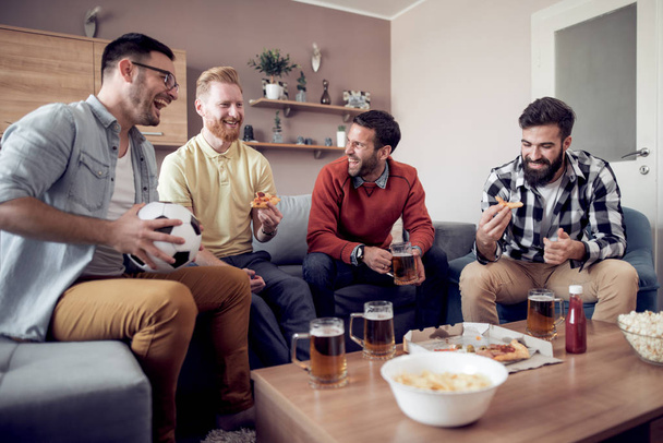Four male friends watching TV at home while eating pizza and drinking beer. - Фото, изображение