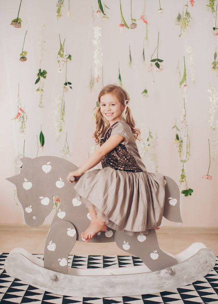 niedliches kleines Mädchen im Kleid mit Spielzeugpferd zu Hause, glückliche Kindheit Konzept  - Foto, Bild